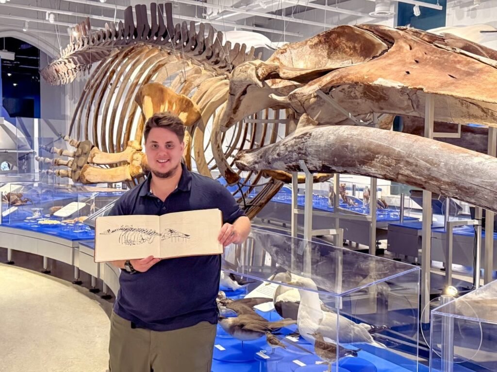 Urban sketcher Owen Labbé holds his sketchbook beside whale skeletons in a natural history museum