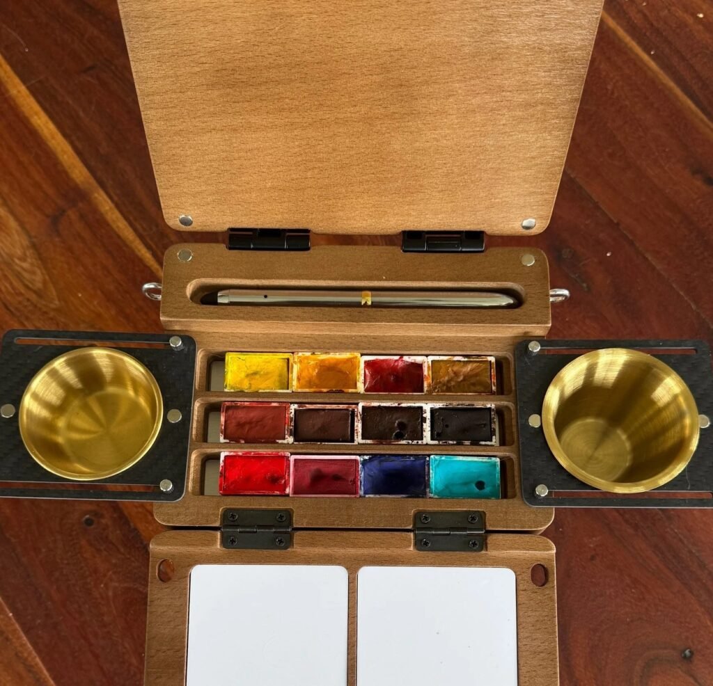 Photo showing an open Fuumuui watercolour travel wooden palette with paints , brass water containers and travel brush