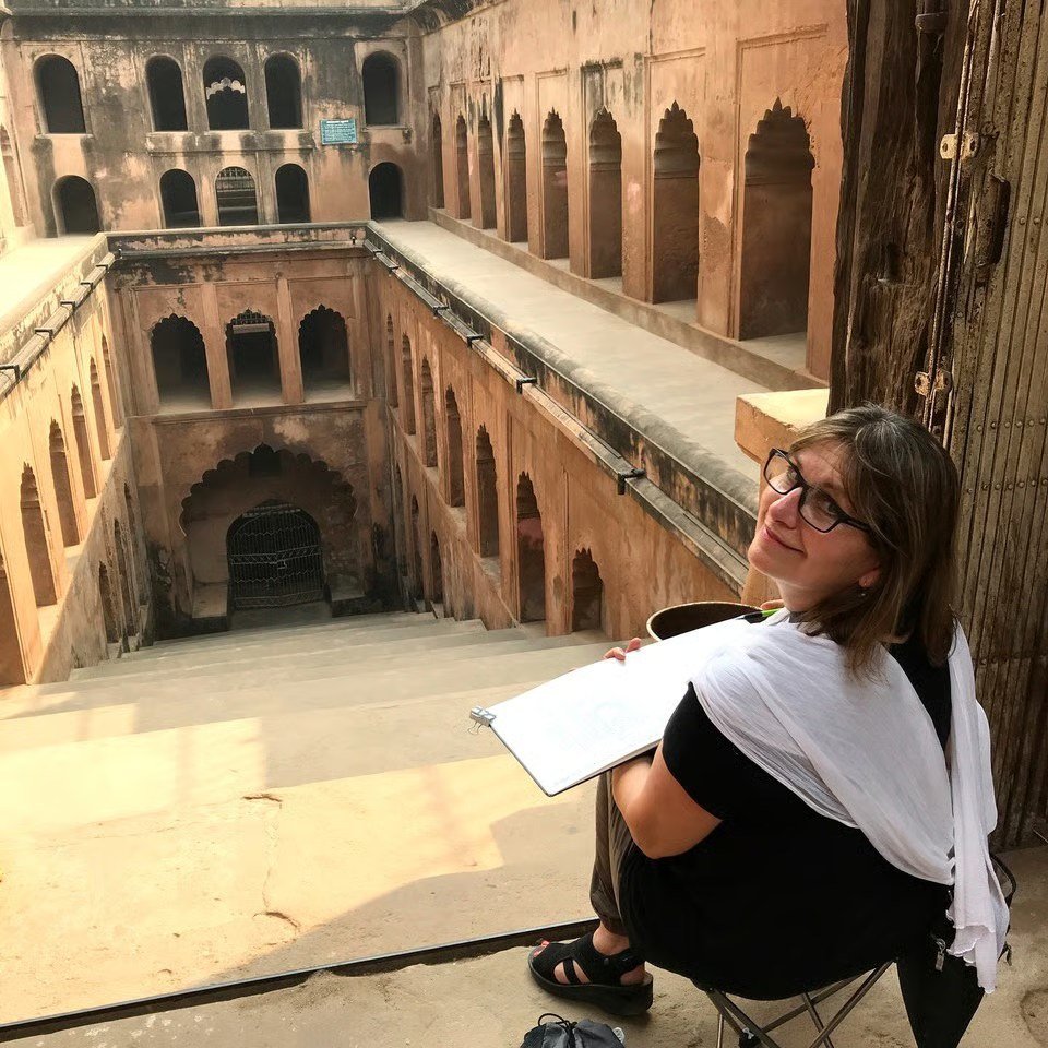 Photo of Stephanie Bower artist holding a sketchbook and looking towards the camera in front of the scene she is sketching